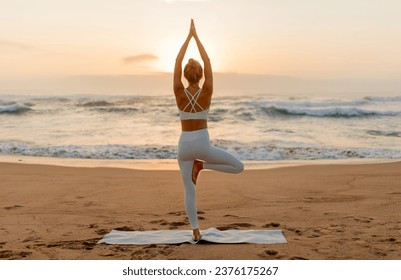 Woman in white activewear strikes a balanced yoga pose, with the gentle sunset illuminating the waves and the sandy beach behind her, back view, full length - Powered by Shutterstock