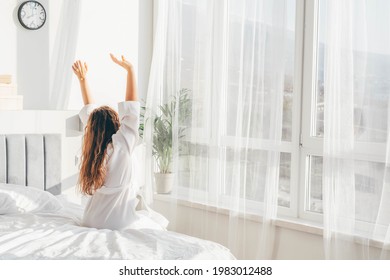  Frau in weißem Bademantel, die sich nach dem Aufwachen in der Nähe des großen weißen Fensters auf dem Bett befindet. 