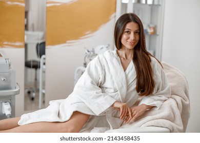 Woman in white bathrobe smiling at camera, lying on daybed in spa center. Laser epilation concept