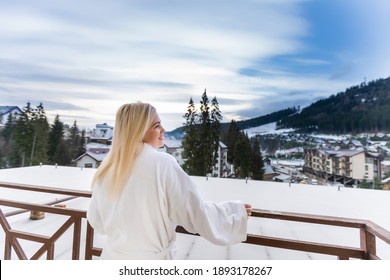 A Woman In A White Bathrobe Standing On The Balcony Of A SPA In Austria In Winter. She Has Wet Hair. She Is Enjoying The View On Mountains In Front Of Her. She Is Feeling Relaxed