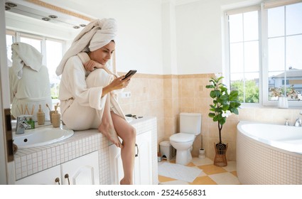 A woman in a white bathrobe with a towel on her head sits on a bathroom counter using her smartphone. The bright, modern bathroom has large windows, a bathtub, plants, and neutral tones. - Powered by Shutterstock