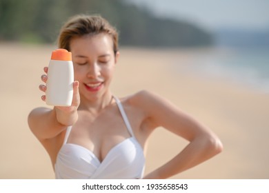 Woman In White Bikini Holding Sunscreen Bottle In Hand On The Beach.