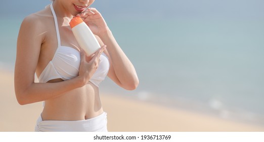 Woman In White Bikini Holding Sunscreen Bottle In Hand On The Beach.