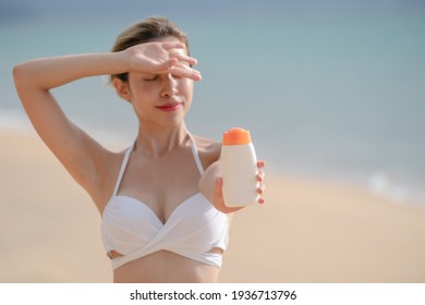 Woman In White Bikini Holding Sunscreen Bottle In Hand On The Beach.