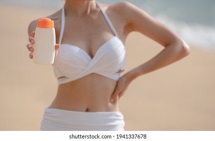 Woman In White Bikini Holding Sunscreen Bottle In Hand On The Beach.