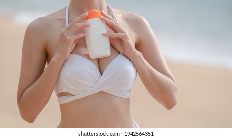 Woman In White Bikini Holding Sunscreen Bottle In Hand On The Beach.