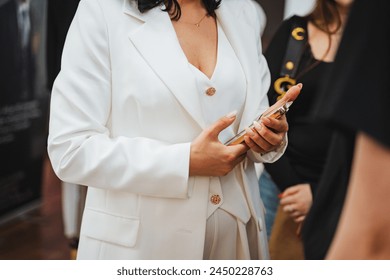 A woman in a white blazer is elegantly holding a cell phone with her thumb on the screen. Her nail polish complements her formal attire, creating a chic and stylish look for the event - Powered by Shutterstock