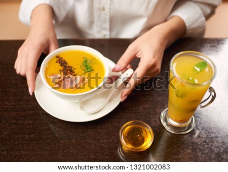 Woman in white blouse ordering tasty and delicious meal at restaurant. Girl hands touching white bowl with orange cream soup. Near first dish on brown table standing tea with mint and honey.