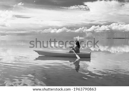 woman in white boat holding paddle oar in still lake water . Romantic creative original expression of dream subconscious. Wind farm at background. Black and white emotional landscape melancholy
