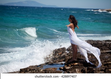Woman in white cape dress stands on the rocky beach on the sea