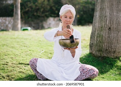 Woman In White Clothes And Cover Hair, Hold Sound Metal Bowl And Make Music, Relation And Meditation Body. Outdoor.  Yoga For Health, Practice For Forgiveness And Spiritual Harmony.