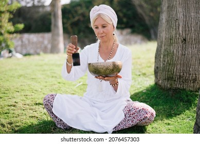 Woman In White Clothes And Cover Hair, Hold Sound Metal Bowl And By Stick Make Music, Relation And Meditation Body. Outdoor.  Yoga For Health, Practice For Forgiveness And Spiritual Harmony.