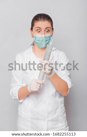 A woman in a white coat, mask and gloves holds a huge syringe in her hands. Portrait of a doctor in uniform on a white background, ready to make an injection.