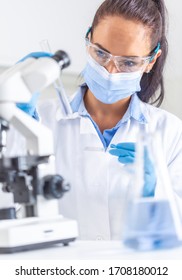 Woman in white coat, protective goggles and face mask looks at test-tube in one hand, holding a microscope slide in the other hand. Coronavirus COVID-19 concept.