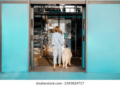Woman with white dog entering supermarket with beautiful turquoise sliding doors. Concept of pet friendly public institutions or shops - Shutterstock ID 2345824717