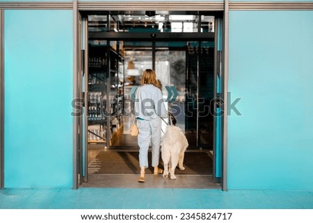 Woman with white dog entering supermarket with beautiful turquoise sliding doors. Concept of pet friendly public institutions or shops