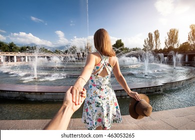 Woman in white dress with hat holding man by hand and going to fountain in the park in Almaty, Kazakhstan