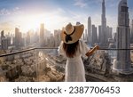 A woman in a white dress and hat looks at the panoramic sunset view of the downtown district skyline of Dubai, UAE