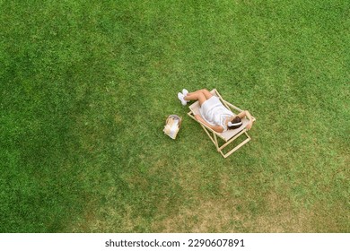 Woman in a white dress in headphones with basket for picnic sitting on deck chair on the green grass sunbathes at summer day. Top view, drone, aerial view.