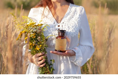 A woman in a white dress is holding a brown glass bottle and a bouquet of herbs. A blank label with space for a description. Alternative Medicine and natural remedies. The idyllic atmosphere - Shutterstock ID 2029759655
