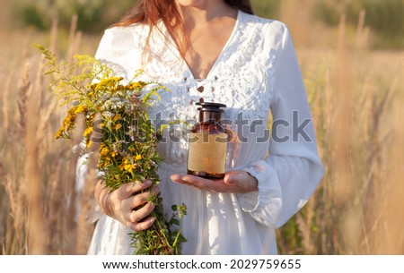 A woman in a white dress is holding a brown glass bottle and a bouquet of herbs. A blank label with space for a description. Alternative Medicine and natural remedies. The idyllic atmosphere