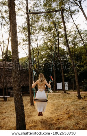 Woman in white dress, with long blonde hair, sitting on swing hanged on trees in the forest.
