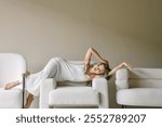 Woman in a white dress lounges elegantly across plush white armchairs, set against a neutral backdrop.