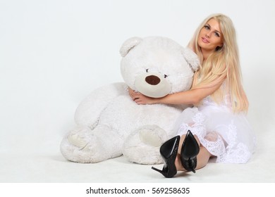 Woman in white dress sits and embraces big toy bear in white studio
