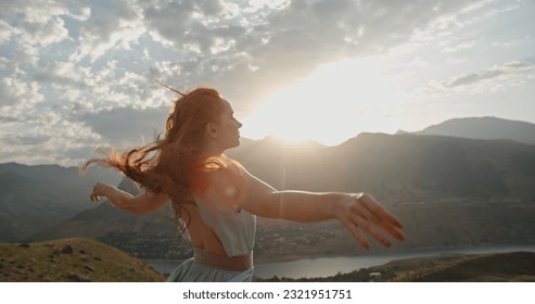 Woman in white dress standing on top of a mountain with raised hands while wind is blowing her dress and red hair - freedom, nature concept . Copy space - Powered by Shutterstock