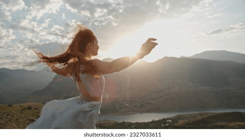 Woman in white dress standing on top of a mountain with raised hands while wind is blowing her dress and red hair - freedom, nature concept - Powered by Shutterstock