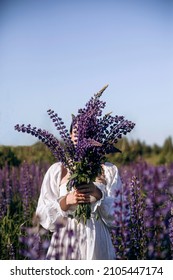a woman in a white dress stands in the middle of the field and holds a bouquet of wildflowers in her hands. girl covers her face with a bouquet of purple lupines. flowering lavender