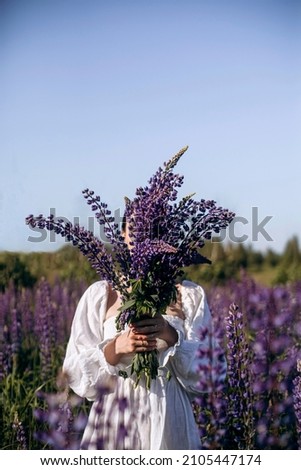 a woman in a white dress stands in the middle of the field and holds a bouquet of wildflowers in her hands. girl covers her face with a bouquet of purple lupines. flowering lavender