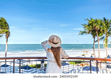 Woman in white dress with straw hat, observing sea view from hotel balcony with palms and beach view. Resort leisure and relaxation. - Powered by Shutterstock