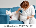 A woman in white gently petting a white Siberian Husky relaxing on a blue sofa, showing a moment of affection and bonding. Perfect for pet and lifestyle themes