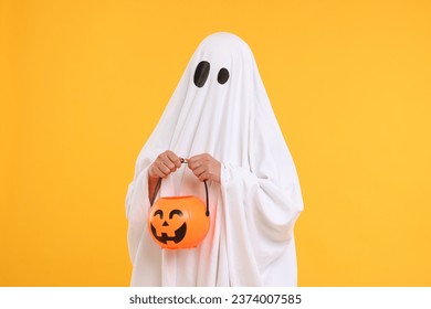 Woman in white ghost costume holding pumpkin bucket on yellow background. Halloween celebration