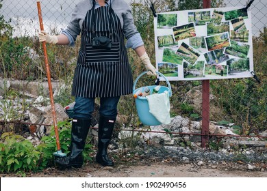 Woman In White Gloves With Cleaning Supplies Near Garbage Dump. Says No To Environment Polution. Poster Parks Instead Of Landfills On A Chain Link Fence