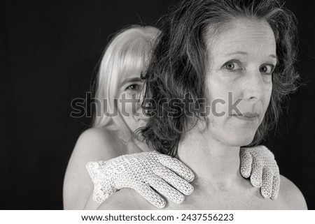 Woman with white hair holds her hand on the neck of a woman with dark hair and they look teasingly into the camera