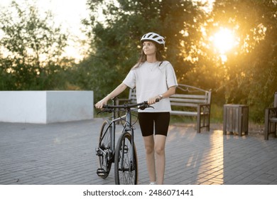 woman with a white helmet walks beside her bicycle on a paved path. She is wearing a white t-shirt, black shorts, and white sneakers - Powered by Shutterstock