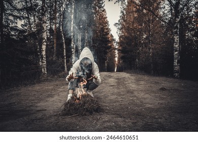 A woman in a white hoodie crouches on a dirt forest path, intensely focused on a small fire she has started. Tall birch trees with lush green foliage line the path, adding to the serene yet intense - Powered by Shutterstock