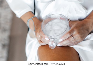 A Woman In A White Housecoat Holds A Glass Of Water With Ice In Her Hands. Cold Water With Ice In A Glass Close-up. Cold Water Is A Way To Cool Off In Hot Weather