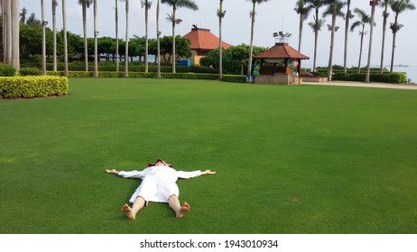 A woman in a white lab coat lies on a green lawn with her arms outstretched, palm trees in the background