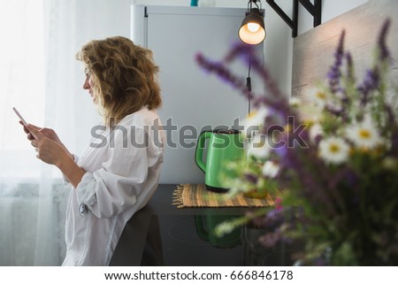 Woman in a white man's shirt is standing in the kitchen, looking at smartphone and smiling. Near the girl there is a bouquet of wild flowers in a vase.