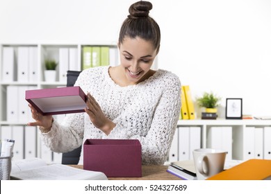 Woman In A White Office Wearing A Sweater Is Happy To See Her Present From A Colleague. Concept Of Secret Santa