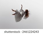 Woman in white ruffled dress floats effortlessly in moment of graceful falling in mid-air against white studio background. Concept of beauty, feminine elegance and purity, dream and reality.