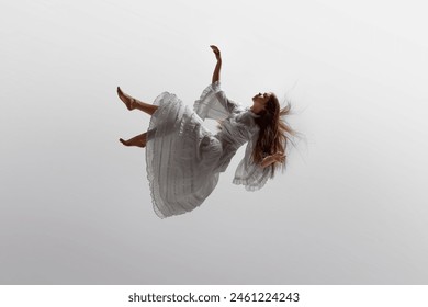 Woman in white ruffled dress floats effortlessly in moment of graceful falling in mid-air against white studio background. Concept of beauty, feminine elegance and purity, dream and reality.
