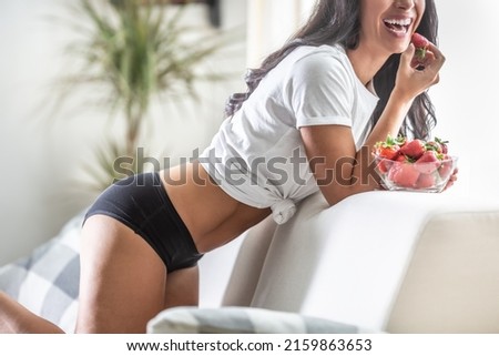 Woman in white shirt and black menstrual panties is laughing and carelessly eating strawberries.