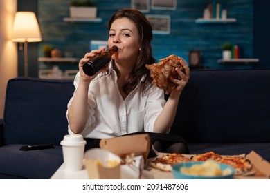 Woman In White Shirt Drinking Beer From Bottle While Holding A Tasty Takeaway Burger Looking At Television Movie. Person Sitting On Couch Eating Delivery Hamburger In Front Of Table With Fast Food.