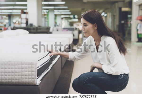 A woman in a white shirt and jeans in a mattress\
store. She examines the mattress she wants to buy. She squats and\
looks at the mattress