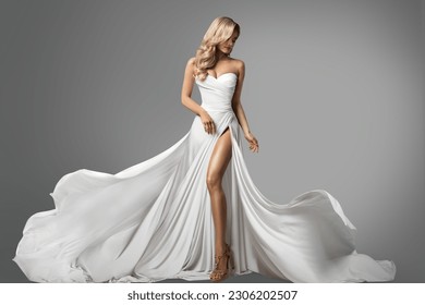 Woman in White Silk Dress with Slit. Beautiful Bride in Wedding Gown Flying on Wind over Gray Background. Fashion Model with Curly Blond Hairstyle