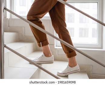 Woman in white sneakers and khaki trousers goes upstairs to her apartment. White staircase in apartment building. Casual outfit, urban fashion. - Shutterstock ID 1981659098